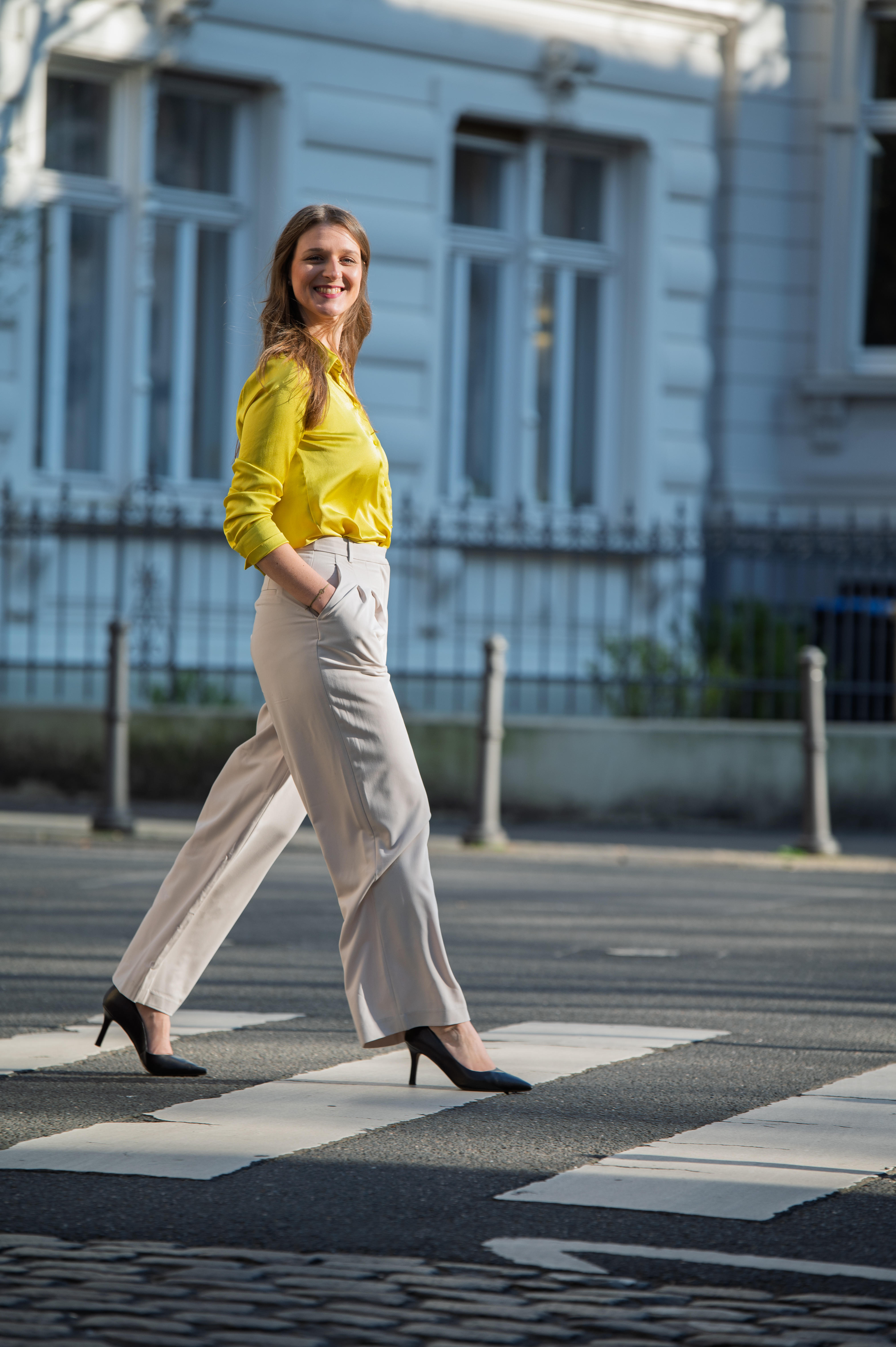 Hier geht eine Business-Frau mit gelber Bluse und Highheels zielstrebig über einen Zebrastreifen und schaut zufrieden in die Kamera.